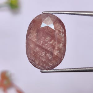 May include: An oval-cut gemstone, held by tweezers. The gem is a muted reddish-brown colour with internal veining and a polished surface. The facets reflect light, creating a sparkling effect. The background is a soft, blurred white.