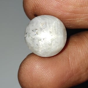 Op de afbeelding: Een close-up van een witte, bolvormige edelsteen met een geometrisch patroon. De edelsteen wordt tussen twee vingers gehouden. Het oppervlak van de edelsteen heeft een kristallijn uiterlijk met donkere insluitsels.