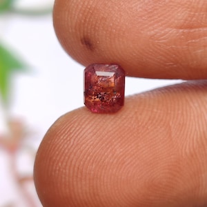 May include: A small, rectangular-cut gemstone with a reddish-brown hue. The gem is held between two fingers, showcasing its facets and internal inclusions. The stone appears to be a tourmaline or similar precious stone.
