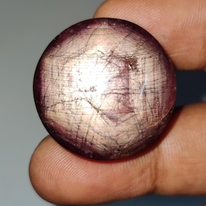Könnte beinhalten: Ein runder, im Cabochon-Schliff gefertigter Sternrubin mit einem tiefen rot-violetten Farbton. Die Oberfläche zeigt einen ausgeprägten sechsstrahligen Sterneffekt, bekannt als Asterismus, der durch Lichtreflexion an internen Einschlüssen entsteht.