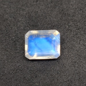 Puede incluir: Una gema rectangular facetada con un brillo iridiscente azul y blanco. La gema se encuentra sobre un fondo oscuro y texturizado, resaltando su claridad y color. El corte de la gema muestra su juego de luces interno.