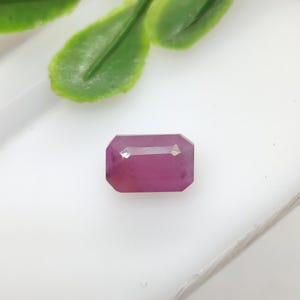 May include: A rectangular, faceted ruby gemstone with a deep pink hue. The gem is set against a white surface, with green leaves in the background. The ruby's cut allows light to reflect, showcasing its clarity and color.