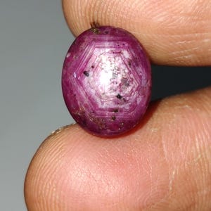 Peut inclure: Pierre précieuse de forme ovale, un rubis. La gemme est d'un rouge profond, avec des motifs hexagonaux internes et des inclusions sombres. La surface polie est mise en valeur, tenue entre les doigts, révélant sa couleur naturelle.