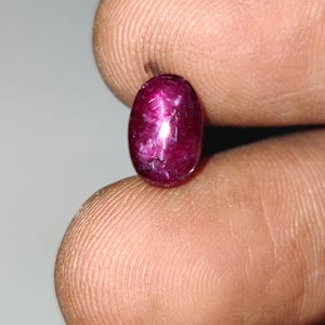 Op de afbeelding: Een diep magenta-kleurige ovale edelsteen met een glad, gepolijst cabochon oppervlak. De steen vertoont een duidelijke stervormige lichtreflectie in het midden, kenmerkend voor asterisme, met zichtbare interne insluitingen en texturen.