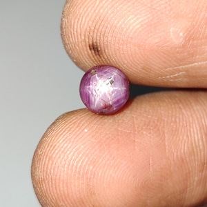 May include: A small, round, cabochon-cut star ruby gemstone with a deep purplish-pink hue. The stone displays a distinct six-rayed asterism effect, characterized by a bright, intersecting white star pattern visible on its polished, domed surface.