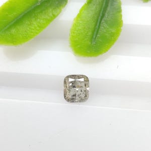 May include: A cushion-cut salt and pepper diamond with a translucent, greyish-brown appearance and visible internal inclusions, showcasing a faceted surface that reflects light across its square-shaped, rounded-corner silhouette.