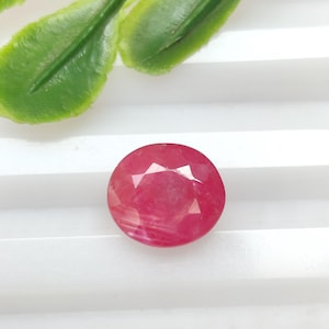 Pode incluir: Uma pedra preciosa de rubi facetada e redonda com um tom vermelho profundo. A pedra preciosa está sobre um fundo branco, com folhas verdes no canto superior esquerdo. O corte do rubi cria múltiplas superfícies refletoras.