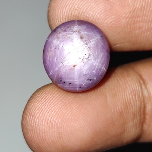 Può includere: Una gemma rotonda e lucida, di colore viola chiaro, con un effetto a stella. La gemma ha una superficie liscia con sottili variazioni di colore e piccole inclusioni. La gemma è tenuta tra due dita.