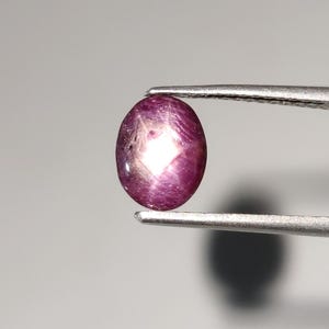 May include: A polished, oval-shaped ruby gemstone, held by tweezers. The gem is a deep reddish-purple color with a star-like effect visible in the center. The background is a soft, neutral gray.