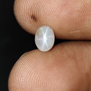 May include: A small, oval-shaped, off-white gemstone with a star-like pattern in the center. The gem is held between two fingers against a dark background. The star effect is created by light reflecting off inclusions within the stone.