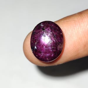 May include: An oval-shaped gemstone with a deep magenta colour. The gem has a reflective surface with internal inclusions, held between two fingers against a white background. The stone's colour varies from dark purple to a lighter pink.