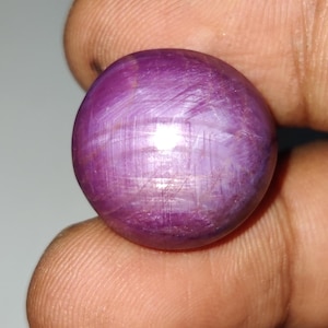 May include: A round, cabochon-cut star ruby gemstone with a deep magenta-purple hue. The surface displays a distinct six-rayed asterism effect, characterised by intersecting light lines that create a star pattern across the polished, translucent stone.