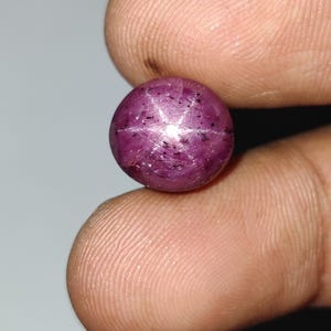 Puede incluir: Una gema de rubí estrella, pulida y de forma redonda. La gema es de color rojo púrpura intenso, con un patrón de estrella de seis rayos visible en su superficie. El efecto de estrella se produce por la luz que refleja en las inclusiones.