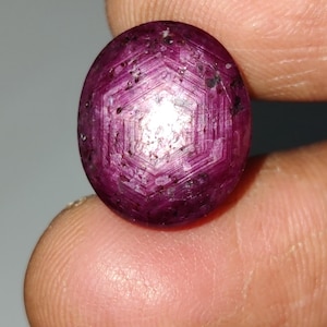 Könnte beinhalten: Ovale, tiefroter Edelstein mit geometrischem Muster. Der Edelstein hat eine polierte Oberfläche und eine komplexe innere Struktur mit sichtbaren Einschlüssen. Der Stein wird zwischen Fingern gehalten, um seine Größe und Farbe zu zeigen.