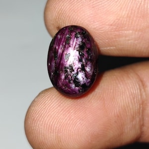 May include: An oval gemstone in a deep purple colour, featuring black and white inclusions. The polished surface reflects light, highlighting the stone's internal structure. Held between fingers, the gem's size is apparent.