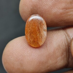 May include: Oval-shaped gemstone with a warm orange hue, featuring internal patterns of lighter shades. The polished surface reflects light, showcasing the stone's natural inclusions. The gem is held between fingers, highlighting its size and shape.
