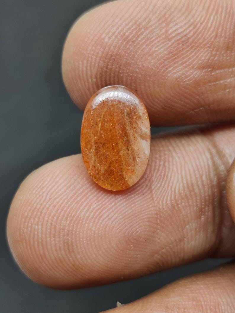 May include: Oval-shaped gemstone with a warm orange hue, featuring internal patterns of lighter shades. The polished surface reflects light, showcasing the stone's natural inclusions. The gem is held between fingers, highlighting its size and shape.