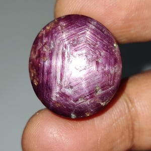 Puede incluir: Una gema redonda y pulida de color magenta intenso. La piedra presenta un patrón en forma de estrella en su superficie, con líneas visibles y un brillo sutil. La gema se sostiene entre los dedos, mostrando su tamaño y forma.