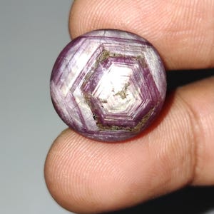 May include: A round, faceted gemstone with a complex geometric pattern. The gem features shades of purple and white, with a central hexagonal design. The stone is held between fingers, showcasing its polished surface and intricate internal structure.
