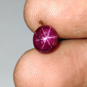 Op de afbeelding: Een ronde, cabochon geslepen sterrobijn edelsteen met een diepe magenta-rode kleur. De steen vertoont een duidelijk zesstralig stereffect, bekend als asterisme, dat verschijnt als een heldere witte lichtreflectie in het midden.