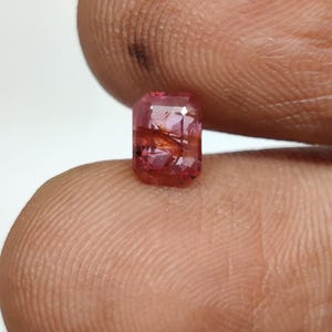 Könnte beinhalten: Ein rechteckiger facettierter Edelstein mit einem leuchtend rosa und rötlichen Farbton, der durch die transparente Oberfläche sichtbare nadelartige Einschlüsse aufweist. Der Stein zeigt einen klaren Schliff mit mehreren Facetten, die das Licht reflektieren.