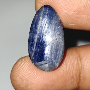 Könnte beinhalten: Ein polierter, ovaler Edelstein in tiefem Blau mit helleren blauen und grauen Streifen. Zwischen Fingern gehalten, reflektiert die glatte Oberfläche des Steins das Licht. Die Tropfenform ist deutlich. Ein schöner Stein.