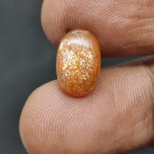 May include: Oval-shaped sunstone gemstone with a shimmering, golden-orange color. The polished stone displays a sparkling effect, held between fingers against a dark background. The gemstone is a natural, polished stone.