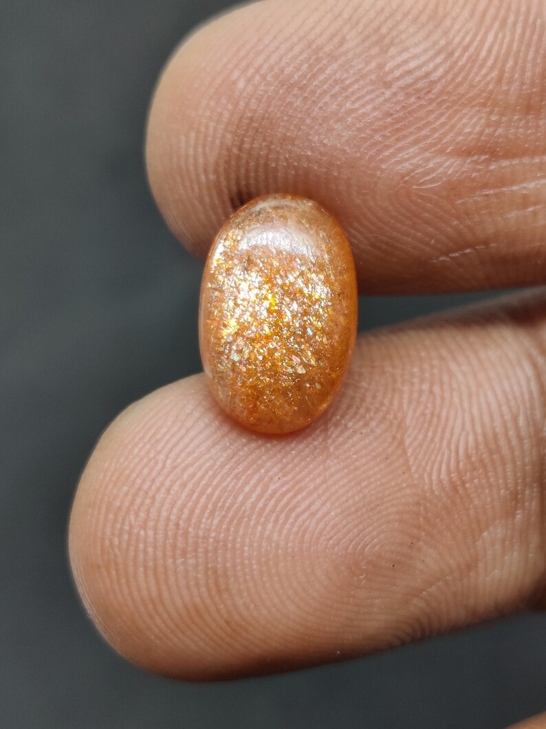 May include: Oval-shaped sunstone gemstone with a shimmering, golden-orange color. The polished stone displays a sparkling effect, held between fingers against a dark background. The gemstone is a natural, polished stone.