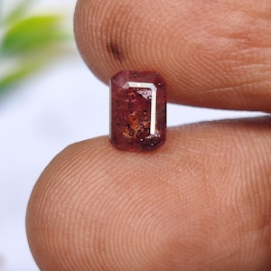 May include: A small, rectangular gemstone with a reddish-brown hue. The gem has a polished surface and a visible internal structure. A white, L-shaped facet is visible on the top right corner. The gem is held between two fingers.