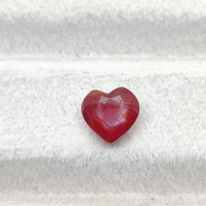 Puede incluir: Una piedra preciosa en forma de corazón de color rojo intenso con una superficie facetada. La piedra presenta una base puntiaguda y una parte superior hendida, mostrando un acabado pulido que refleja la luz a través de sus múltiples planos.