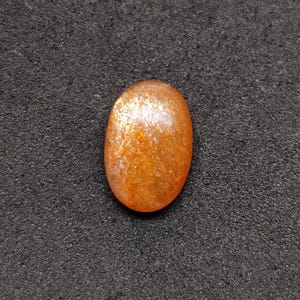 May include: Oval-shaped sunstone gemstone with a shimmering, orange and gold appearance. The polished stone has a smooth surface and is set against a dark, textured background. This gemstone is suitable for jewelry making.
