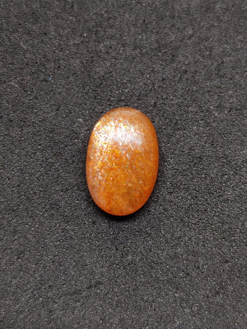May include: Oval-shaped sunstone gemstone with a shimmering, orange and gold appearance. The polished stone has a smooth surface and is set against a dark, textured background. This gemstone is suitable for jewelry making.