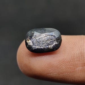 May include: A dark, faceted gemstone with dark blue and silver inclusions. The gem is held between fingers, showcasing its polished surface and unique internal patterns. The stone has a rectangular shape with rounded corners.