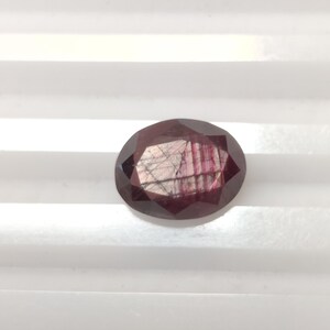 May include: Oval-cut, faceted gemstone with a deep burgundy color. The gem has internal striations and a polished surface, reflecting light. The stone is set against a white, ridged background, highlighting its color and shape. A gemstone for jewelry making.