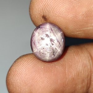 Peut inclure: Une pierre précieuse rubis étoilé de taille cabochon ronde, présentant une teinte translucide violet clair et rose. La pierre affiche un motif distinct en étoile à six branches centré, appelé astérisme, avec des lignes de croissance.