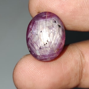 Pode incluir: Uma gema redonda de cor rosa com um padrão em forma de estrela e inclusões pretas. A gema está engastada em um anel de prata.