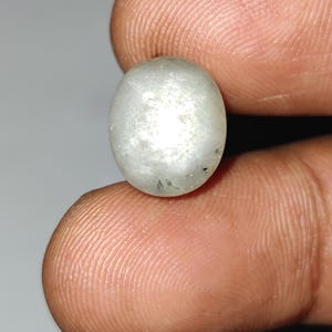 May include: A small, oval-shaped, off-white gemstone. The stone has a slightly translucent appearance with subtle internal inclusions. The gem is held between two fingers, showcasing its size and shape. The overall color is a muted, pale white.