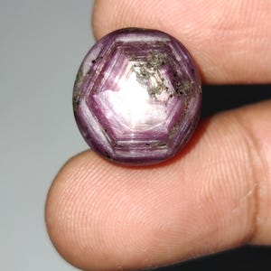 Puede incluir: Una gema facetada y redonda con un tono magenta intenso. La superficie de la gema presenta un patrón geométrico de planos que se cruzan, creando un efecto de estallido estelar. La piedra tiene un aspecto pulido, con áreas de reflexión de la luz.