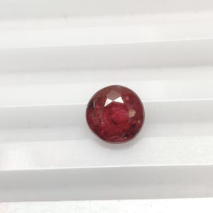 Puede incluir: Una gema facetada, redonda, de color rojo intenso. La gema tiene una forma ligeramente irregular y una superficie pulida. La piedra se encuentra sobre un fondo blanco, resaltando su color y claridad. Esta gema podría usarse en joyería.