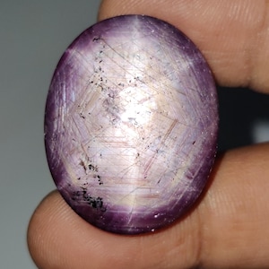 May include: Oval-shaped, polished gemstone with a star effect. The gem features a deep purple hue with lighter, almost white, internal lines creating a starburst pattern. Small black inclusions are visible.