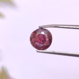 May include: A faceted, oval gemstone held by tweezers. The gem is a deep red colour with lighter internal inclusions. The tweezers are silver, and the background is a soft, blurred white.