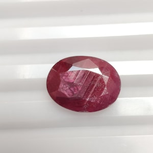 May include: A deep red oval-shaped gemstone with a star-like pattern inside. The gemstone is on a white surface.