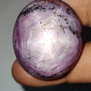 May include: A polished, round gemstone with a lavender hue and subtle white veining. The surface is speckled with small, dark inclusions, creating a unique pattern. The gemstone is held in a hand, showcasing its smooth, spherical shape.