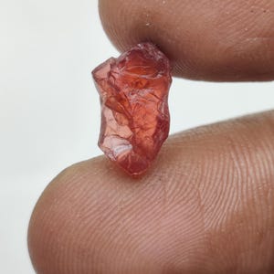 Könnte beinhalten: Ein roher, roter, durchscheinender Edelstein mit einer rauen, unregelmäßigen Form. Der Edelstein wird in einer Hand gehalten.