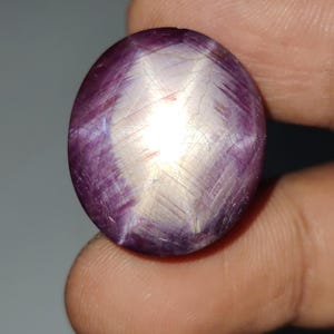 May include: A polished, oval-shaped gemstone with a star effect. The gem features a deep purple hue with a bright, white star pattern radiating from the center. The surface has subtle lines and a slightly textured appearance.