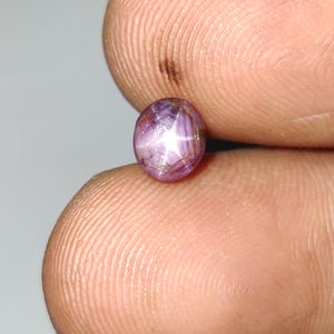 Könnte beinhalten: Ovaler, purpurrosa Edelstein mit einem sechsstrahligen Sternmuster. Der Stein wird zwischen zwei Fingern gehalten, wodurch seine polierte Oberfläche und das innere Lichtspiel hervorgehoben werden. Die Farbe des Steins verändert sich mit dem Licht und erzeugt einen fesselnden visuellen Effekt.