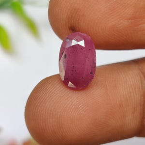 May include: Oval-shaped, faceted ruby gemstone. The ruby is a deep pinkish-red color with small inclusions. The gemstone is held between fingers, showcasing its polished surfaces and clarity. The gem is a focal point, highlighting its natural beauty.