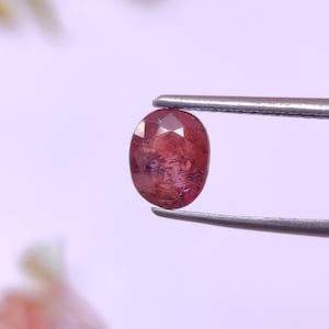 以下が含まれることがあります： ピンセットで保持された、ファセット加工された楕円形の宝石。宝石は、より暗いインクルージョンを持つ深い赤茶色です。ファセットは光を捉え、きらめく効果を生み出します。背景は柔らかくぼやけた紫です。