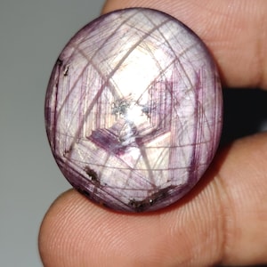 Puede incluir: Una gema redonda y pulida con efecto estrella. La piedra es principalmente de color púrpura claro con líneas que se cruzan creando un patrón de cuadrícula. La gema se sostiene entre dos dedos.