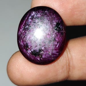 May include: A circular, polished gemstone in a deep purple shade. The stone exhibits a star-like effect with light reflecting from its surface. The gemstone is held between fingers, highlighting its size and colour. The gem has a smooth, rounded form.